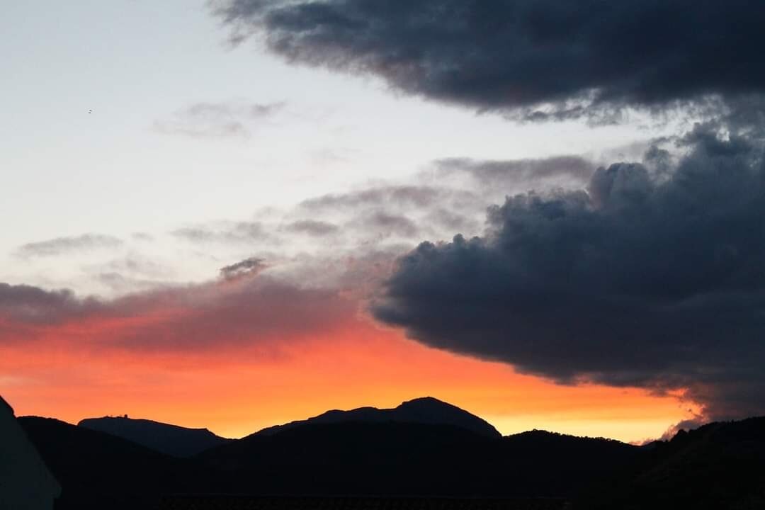 Fotografía del cielo en un atardecer en Alhama de Murcia. En la parte inferior de la foto se ve la silueta de Sierra Espuña. El cielo está dividido por una franja inferior de colores amarillos y naranjas intensos, y por una franja superior de color azul claro. Nubes oscuras al no ser iluminadas, suman texturas a la imagen.