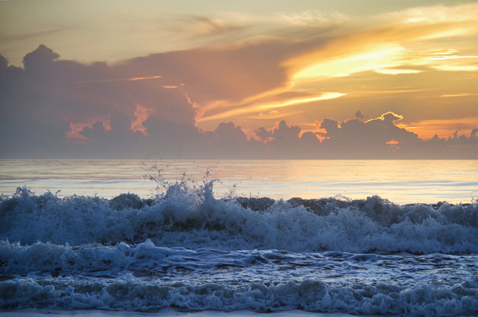 A dramatic ocean scene at sunrise, with golden and purple light spilling through thick clouds. Waves crash in the foreground, their white foam contrasting against the darker water, while the horizon glows softly beneath the colorful sky.