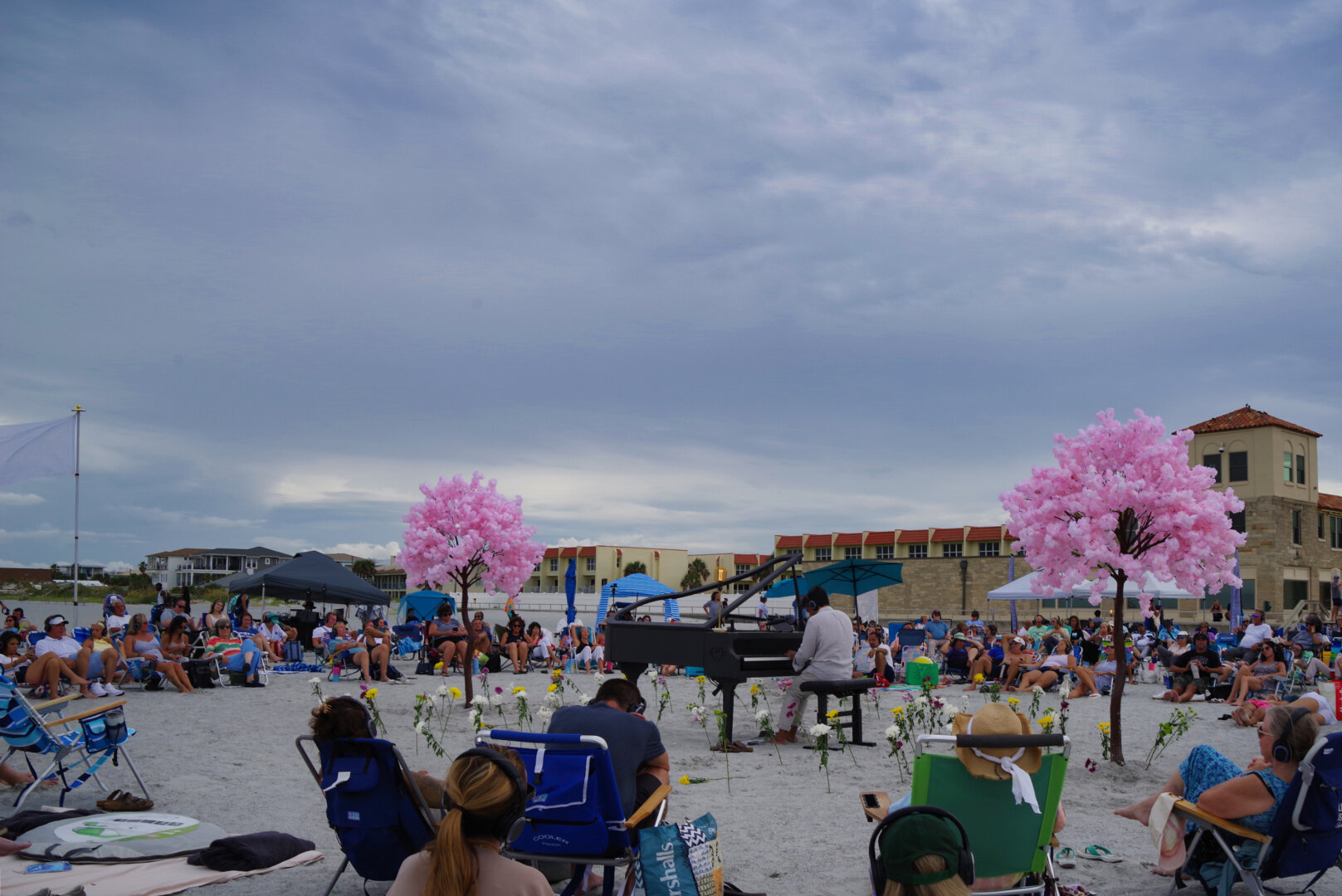 A pianist performs on a grand piano set directly on a sandy beach, surrounded by pink artificial trees and a circular bed of flowers. Dozens of people wearing headphones sit in beach chairs and under umbrellas watching the concert, with red‑roofed buildings and a cloudy sky in the background.