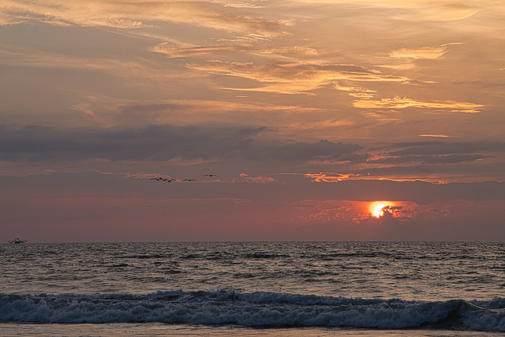 A warm sunrise over the ocean with soft orange and pink light reflecting on gentle waves. A small boat sits near the horizon on the left, and several birds fly above the water, creating a calm, peaceful coastal moment.