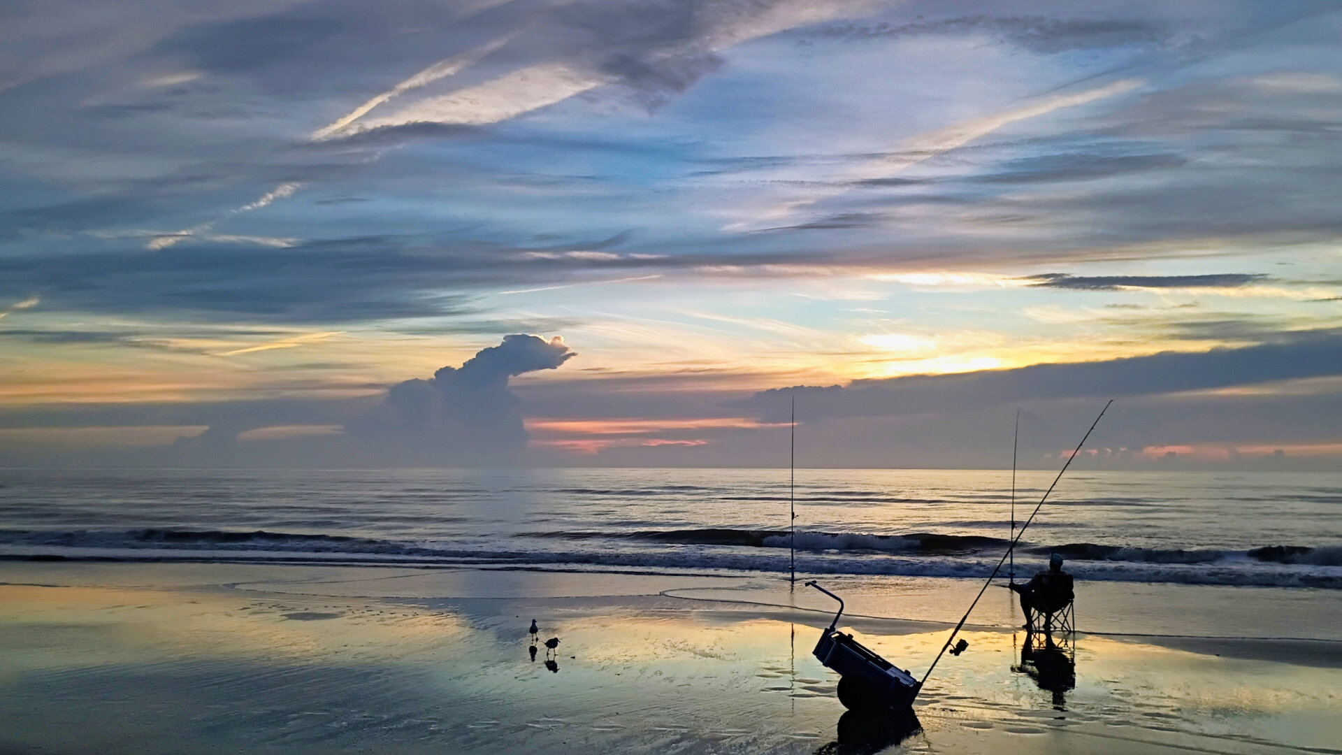 A person sits in a folding chair on a quiet beach at sunrise, fishing with two rods planted in the sand. Soft waves roll toward the shore, reflecting warm orange and yellow light from the low sun behind layered blue‑gray clouds. A small cart or cooler sits nearby, and two birds stand on the wet sand in the foreground.