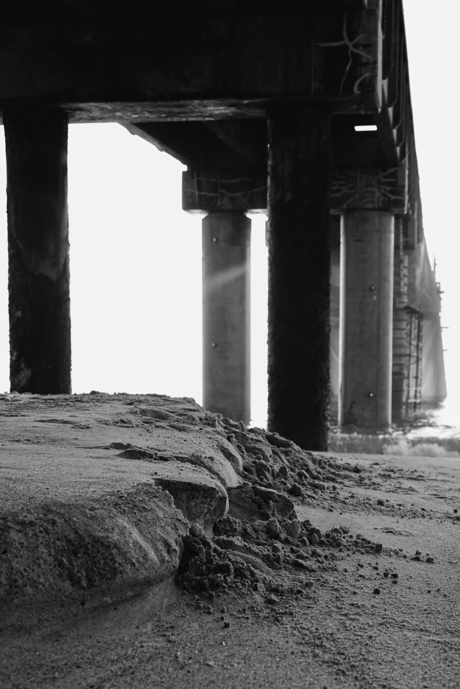 Black‑and‑white view from beneath a pier, showing rough, eroded sand in the foreground and tall concrete pillars receding toward bright light beyond the structure. Strong contrast highlights the geometric lines of the pier and the interplay of shadow and sunlight reflecting off the water.