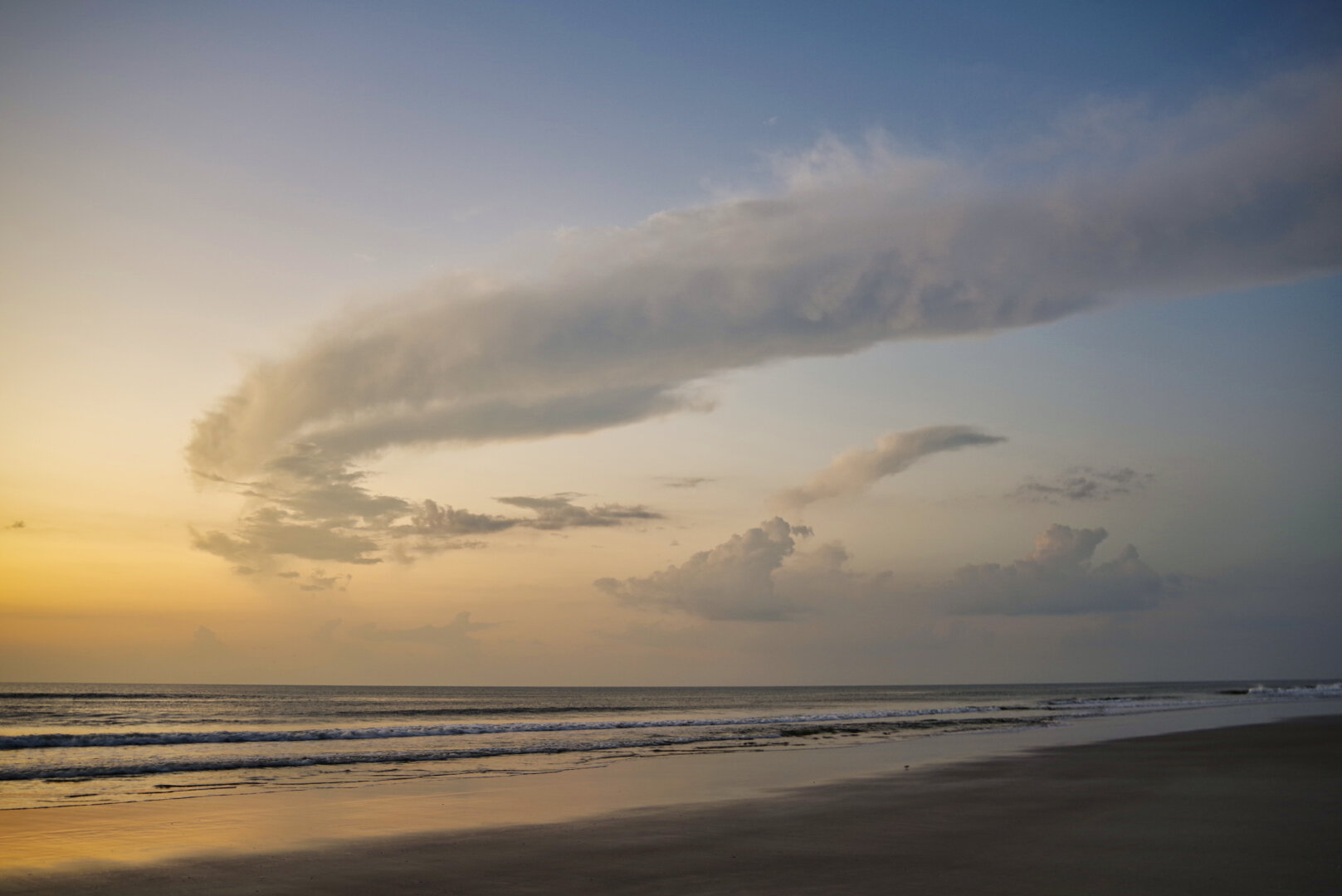 A tranquil beach at sunrise, with gentle waves, wet sand reflecting golden light, and a dramatic sweeping cloud stretching across the sky.