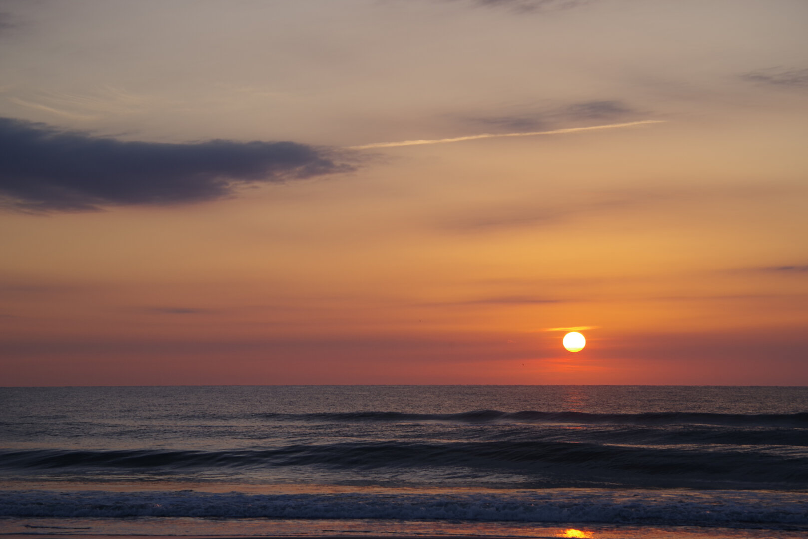 A calm ocean at sunrise, with the sun low on the horizon casting warm orange and pink light across the sky and reflecting on gentle waves rolling toward the shore beneath a few scattered clouds.