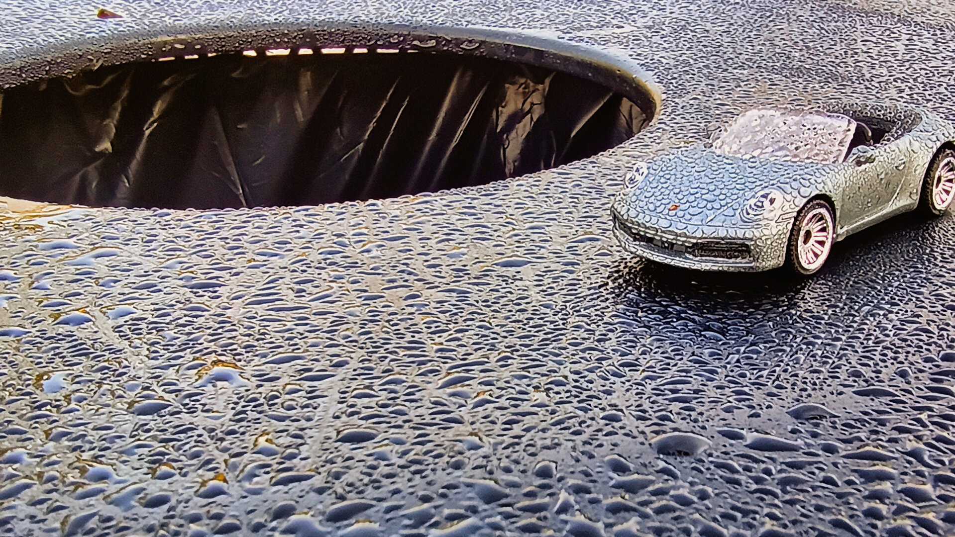 A tiny silver convertible toy car sits on a wet, textured surface covered in glistening water droplets. Its windshield, hood, and headlights are speckled with moisture, and beside it is a circular opening of a trashcan that drops into a dark interior, giving the scene a dramatic, almost cinematic feel despite the miniature scale.