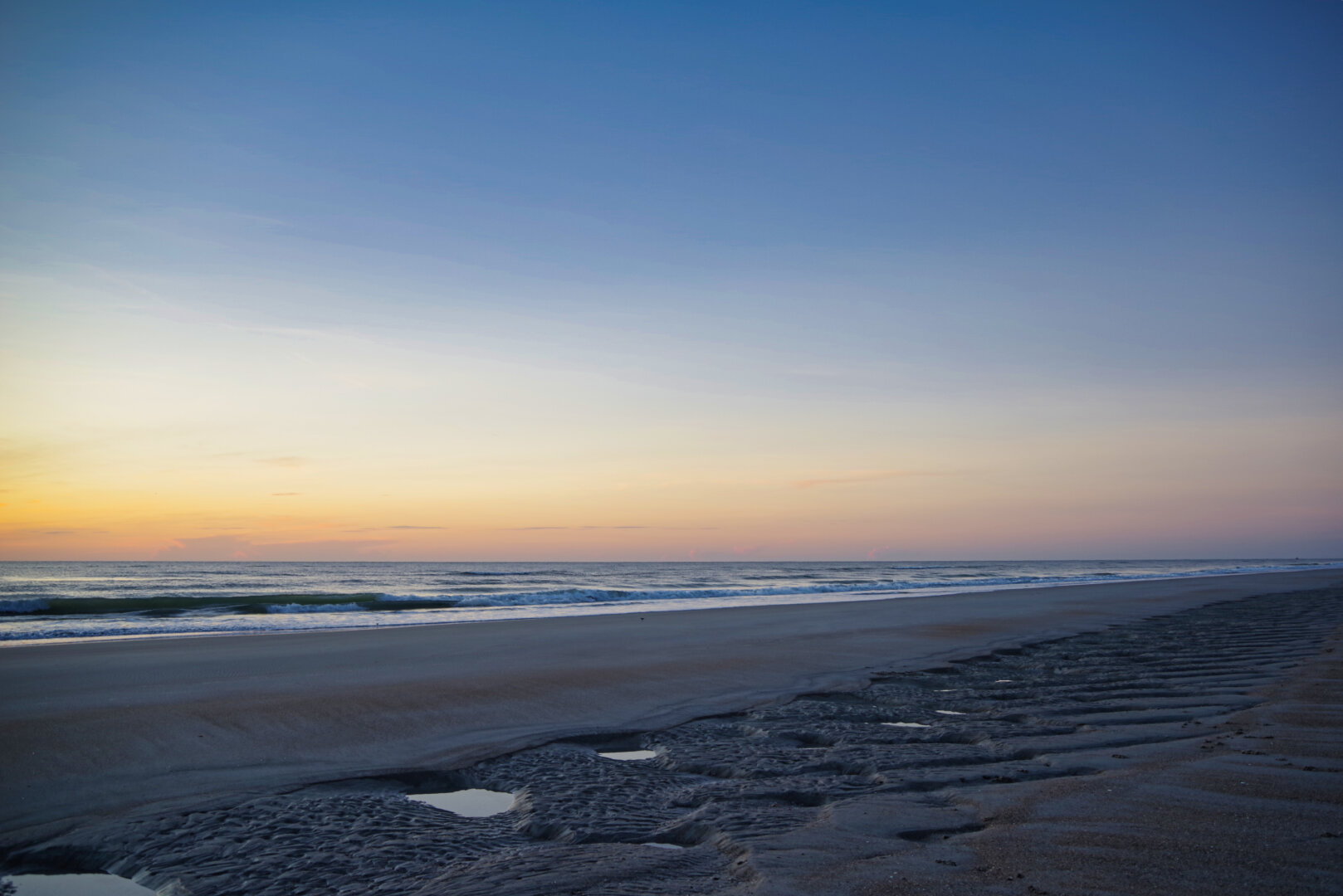 A tranquil beach at sunrise, with rippled wet sand in the foreground reflecting soft light. Gentle waves roll across a calm sea, and the sky fades from warm orange near the horizon to deep blue above.