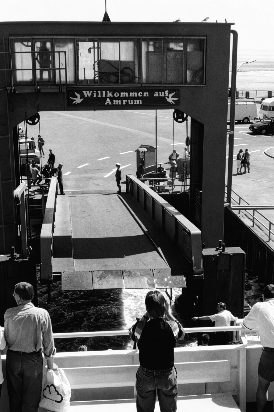 A ferry ramp connects a dock to a white passenger ferry at the Amrum terminal. Several people stand on the dock and on the vessel, watching the loading area. A sign above the structure reads “Willkommen auf Amrum.” Cars and buildings sit in the background under a bright, partly cloudy sky, giving the scene a calm coastal atmosphere.