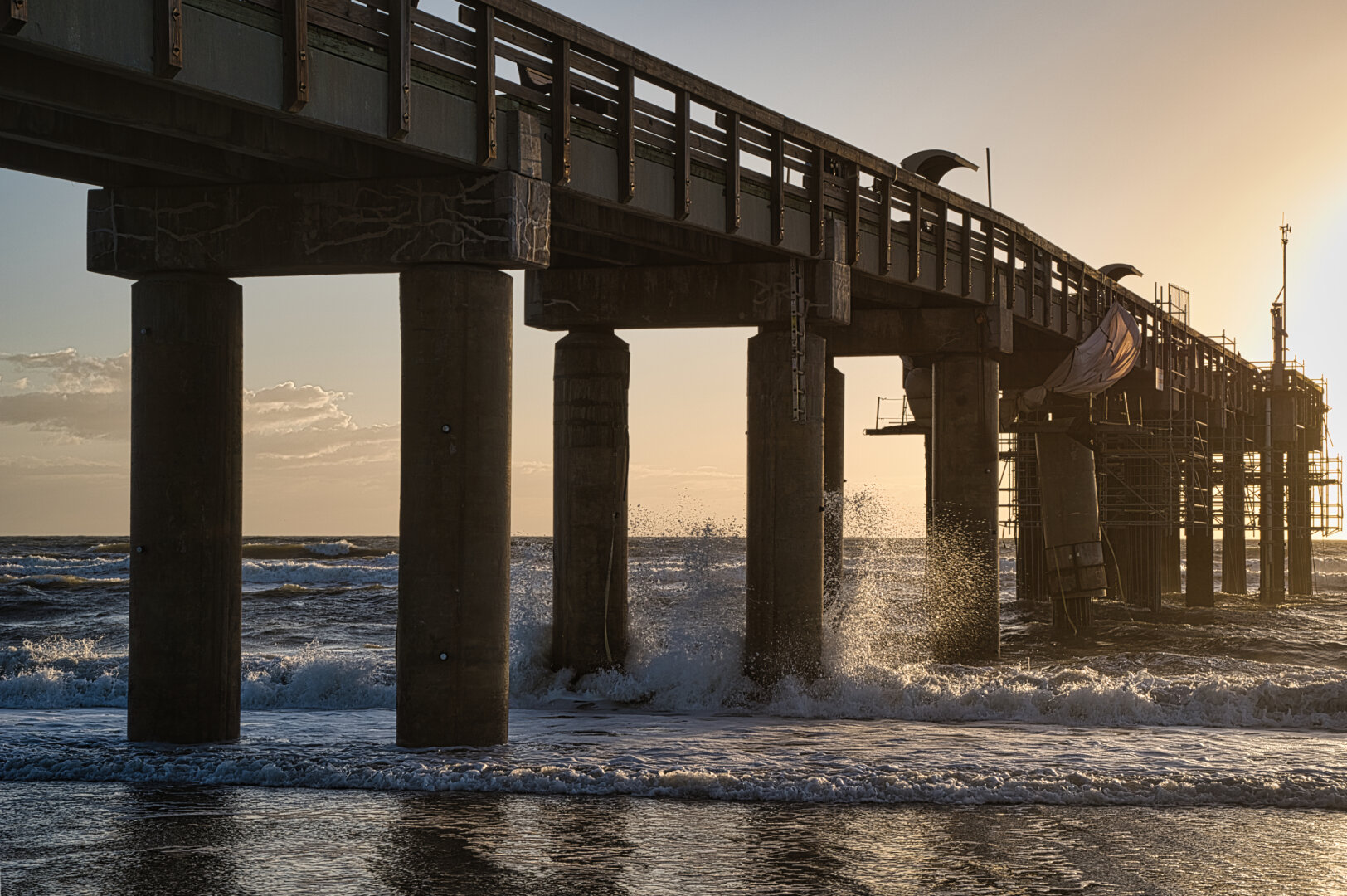 Sunlit ocean waves crash beneath a weathered wooden pier, casting spray and reflections at golden hour in a dramatic coastal scene.