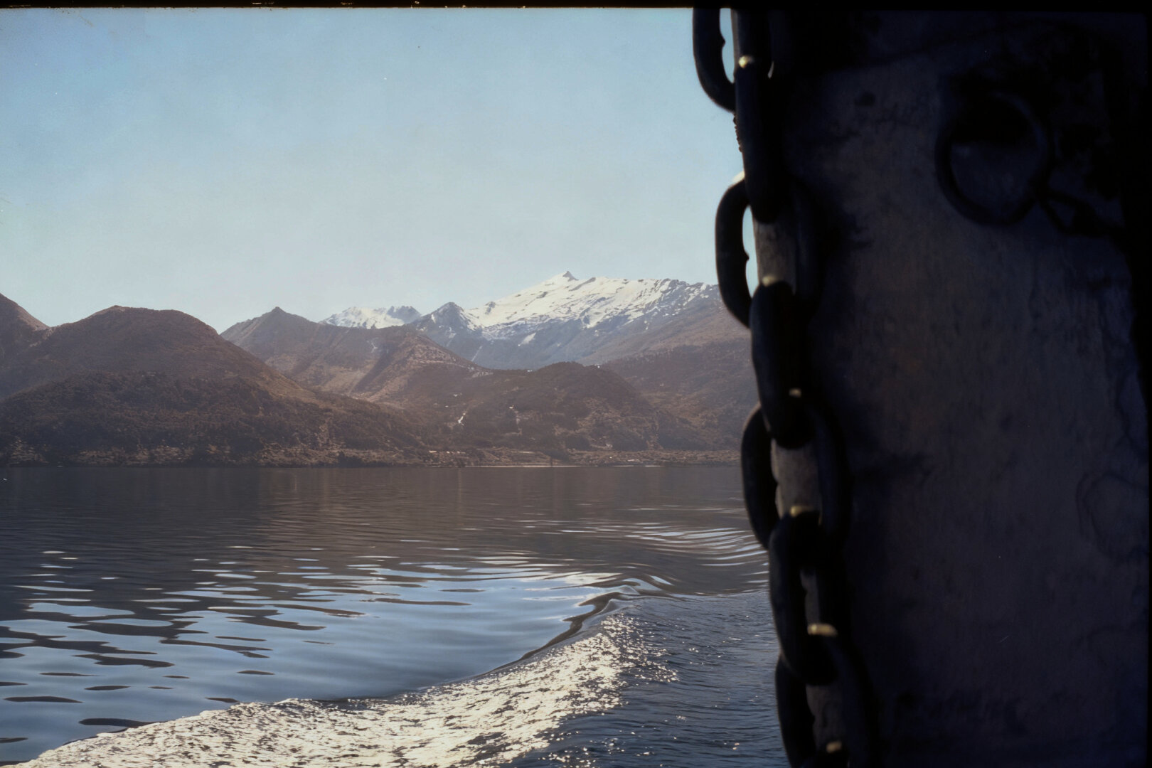A calm mountain lake reflects a range of snow‑capped peaks under a clear sky. Brown, rugged hills sit at the base of the mountains, and a boat’s wake ripples across the water in the foreground. On the right edge of the frame, part of a metal structure with a heavy chain hangs down, indicating the photo was taken from a vessel.