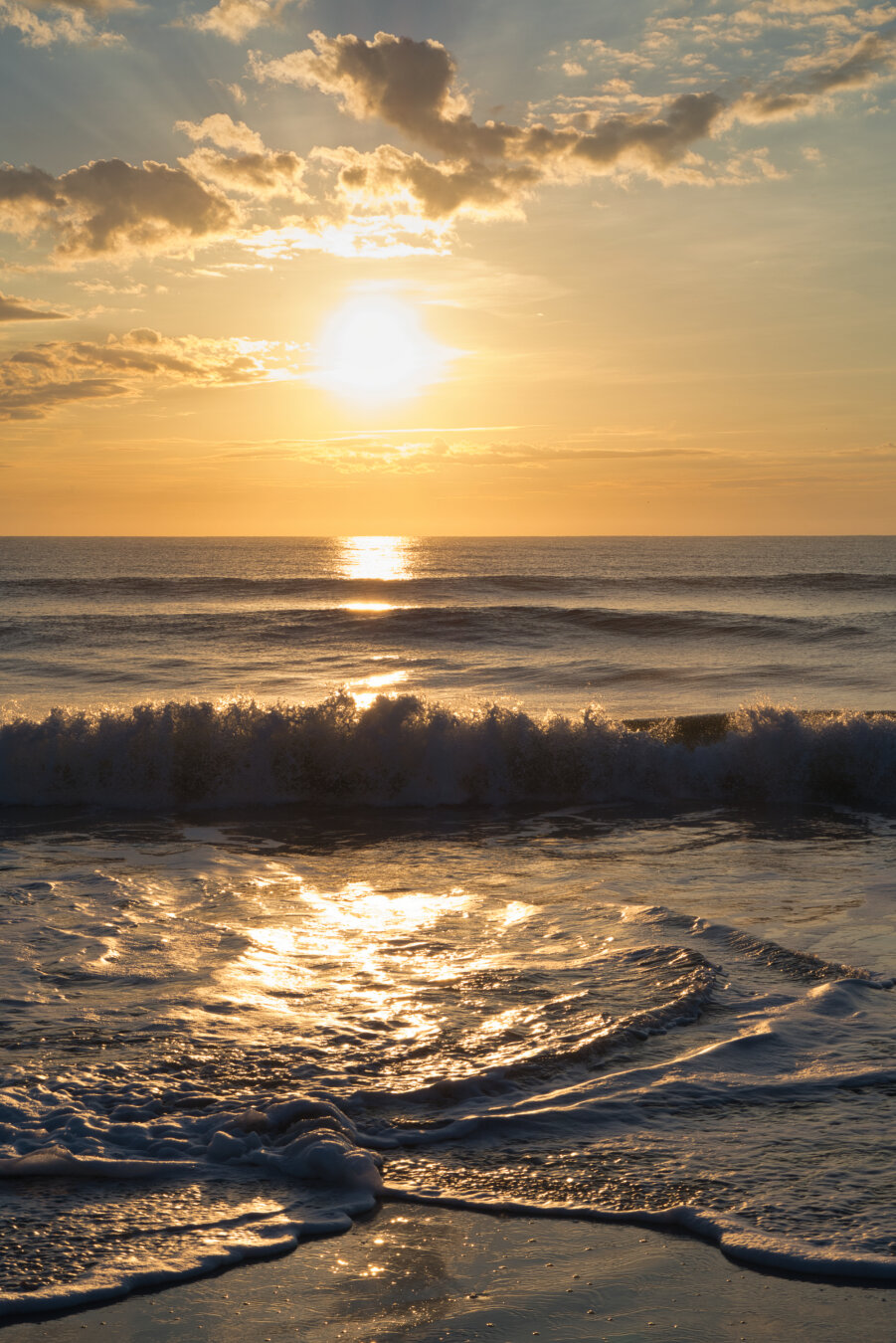 Golden morning light spreads across the ocean as the sun rises at the horizon, tinting the sky with soft amber and pink. Gentle waves roll toward the shore, their white foam catching the early glow, while scattered clouds brighten at the edges in the first light of day.