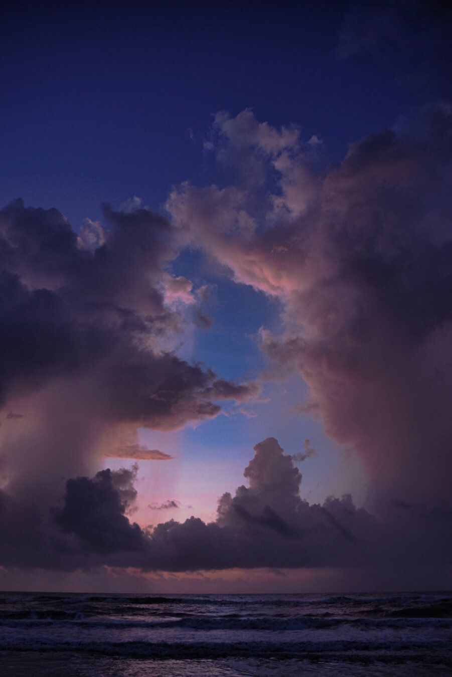 A dramatic sunrise over the ocean, with towering clouds lit in soft pink, purple, and blue hues as early light spreads across the sky. Gentle waves in the foreground catch the pastel colors, creating a vivid contrast between the glowing clouds and the darker water below.
