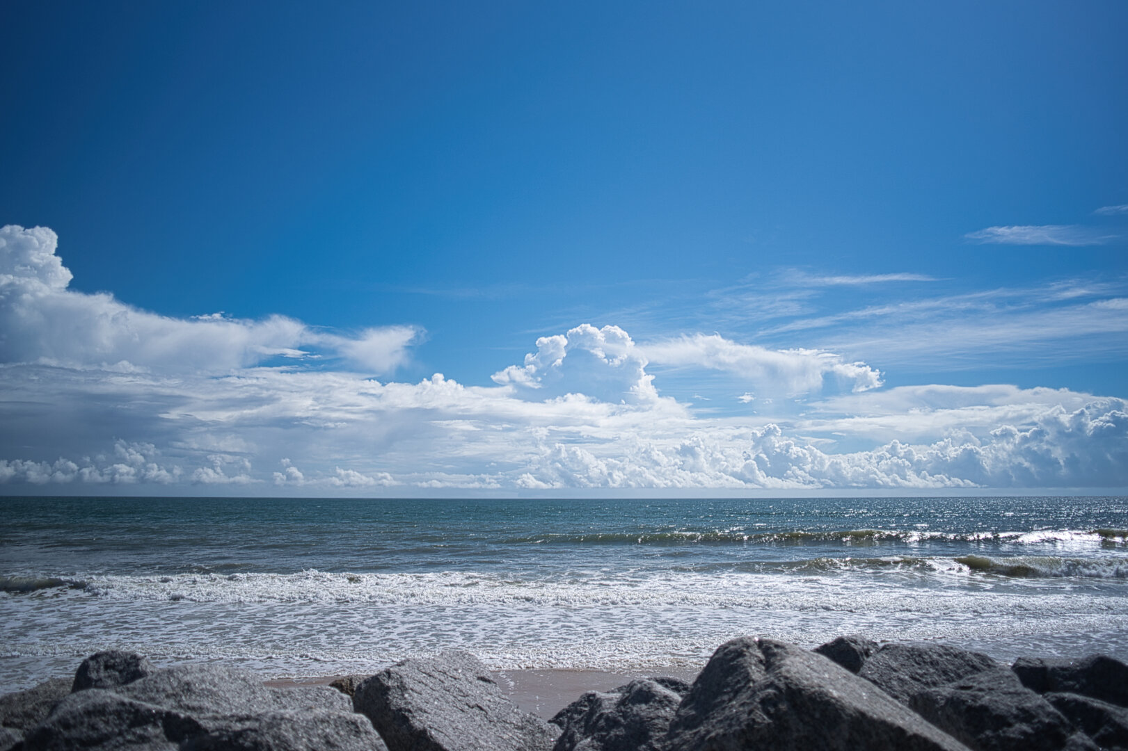 A calm ocean stretches toward the horizon under a bright blue sky dotted with soft white clouds. Gentle waves roll onto a sandy beach, meeting a line of large gray rocks in the foreground. The scene feels open, peaceful, and quietly expansive.