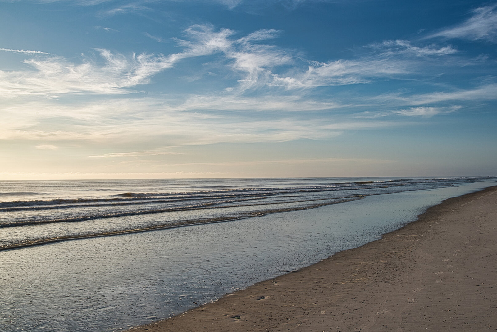 A calm beach with gentle waves washing onto pale sand under a mostly clear sky with soft, wispy clouds. Sunlight reflects on the water, creating a peaceful, open coastal scene.