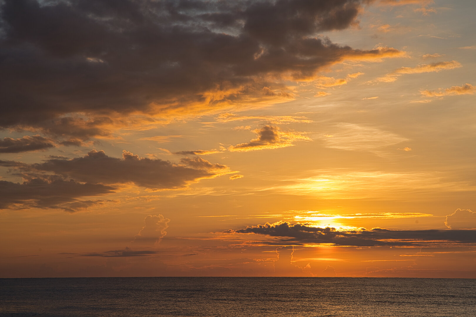 A warm sunrise over a calm ocean, with the sun low on the horizon casting golden and orange light across rippled water. Layers of clouds—some dark, some glowing—stretch across the sky, adding depth and contrast to the scene.