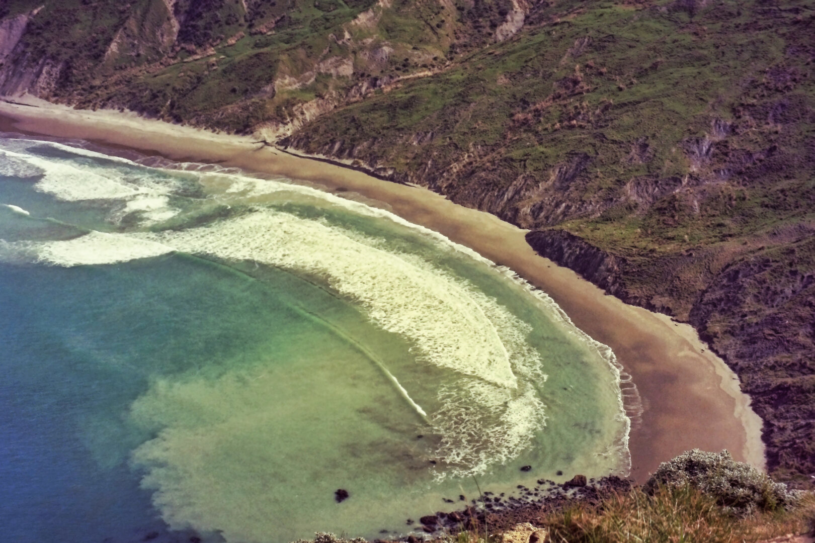 A sweeping aerial view of a rugged coastline where turquoise waves curve into a crescent-shaped sandy beach. Steep, green cliffs rise sharply from the shore, their rocky edges contrasting with the smooth arc of white surf below. The water shifts from deep blue to bright teal as it approaches the beach, highlighting the dramatic meeting of sea and land.