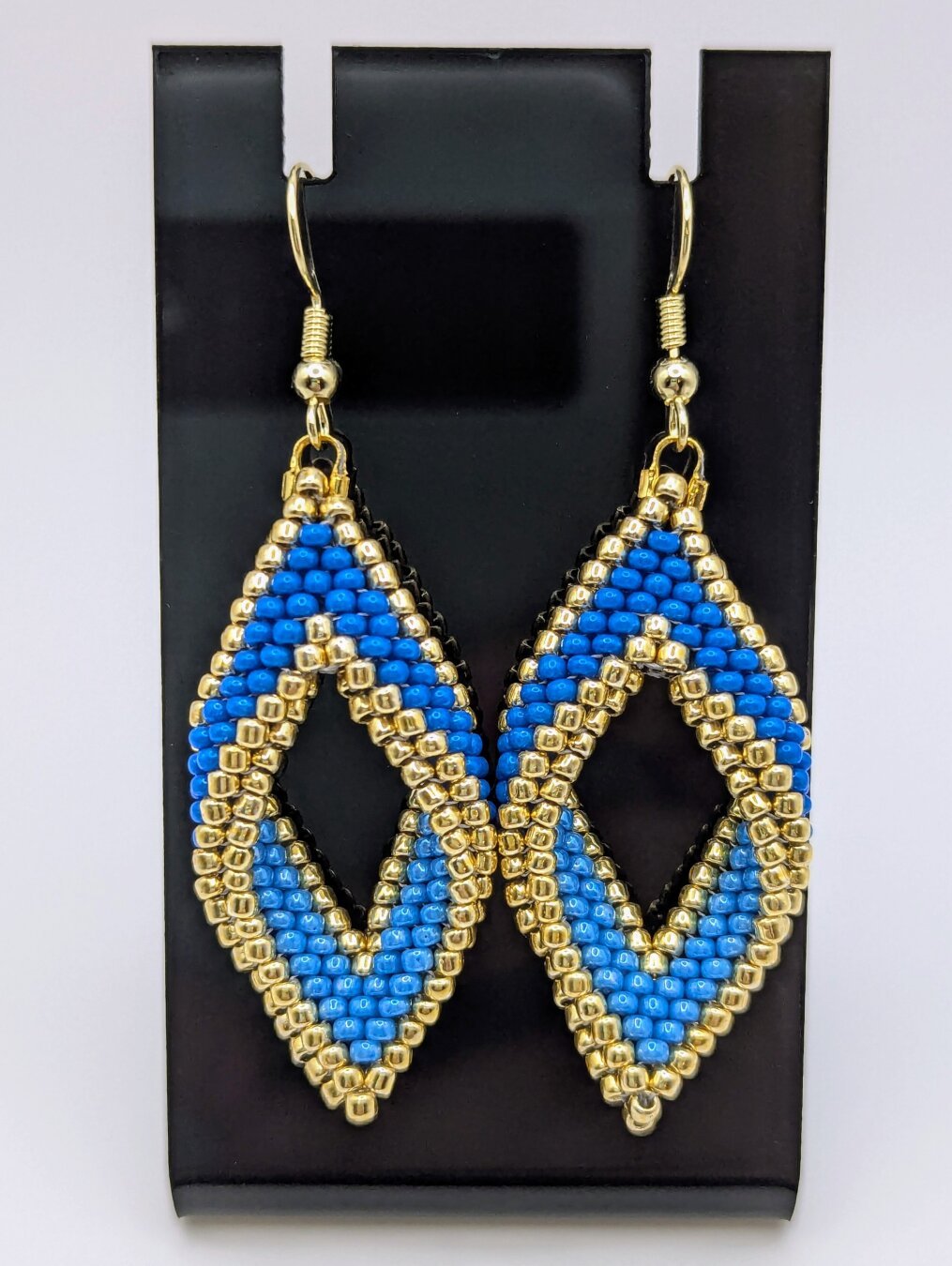 A pair of gold and blue twisted peyote earrings displayed on a shiny black earring stand.