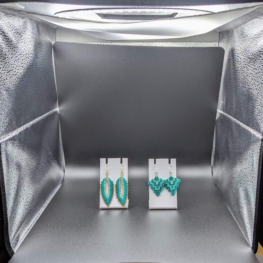 The inside of a small photobox; the current display is a simple black background with two pairs of earrings placed on white stands. The earrings are shades of turquoise and green, and one pair is Russian leaves with gold hooks while the other is peyote arrows with silver hooks.