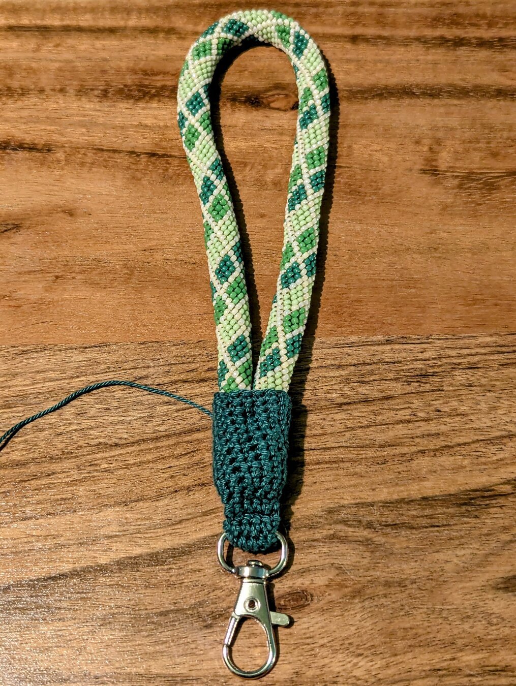 An incomplete diamond patterned bead crochet wristlet lanyard in shades of green and lined with iridescent beige. The silver clip is attached with dark green crochet thread, and the piece isn't finished because the thread part will also be wrapped and secured with beads.