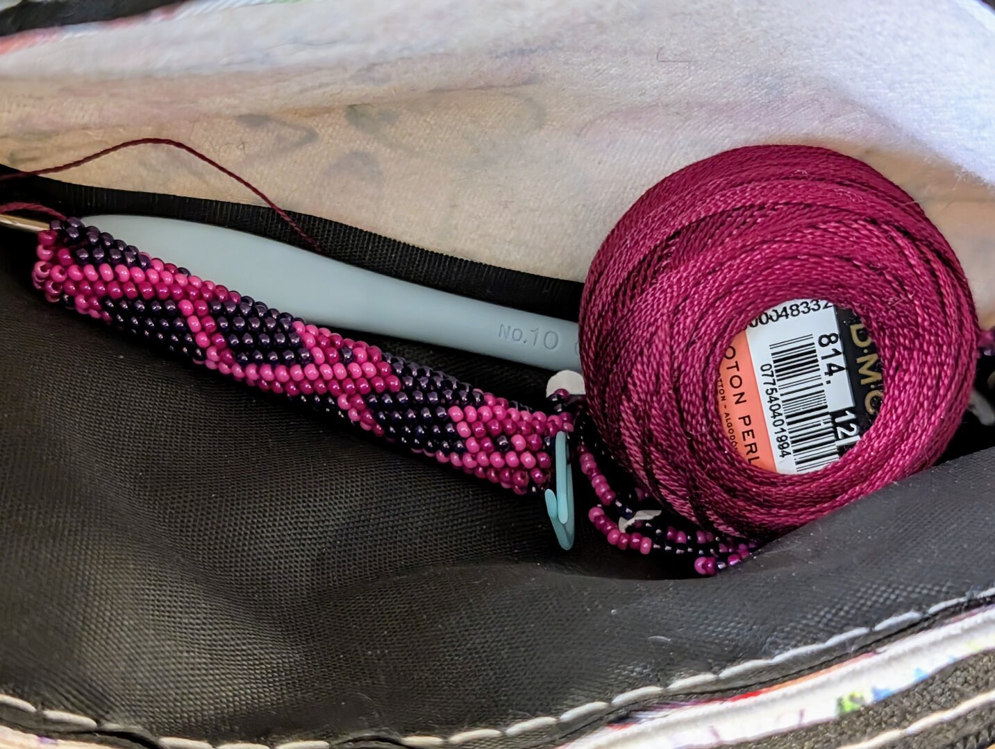 The inside of a pouch that contains bead crochet supplies: burgundy colored cotton crochet thread strung with pink and purple beads, a partially crocheted beaded rope in those colors; the pattern is reminiscent of barbed wire, a blue stitch marker, and a blue hook.