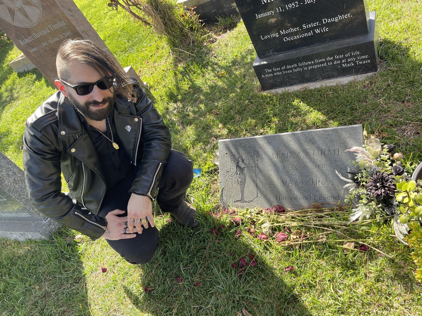 Man, goth style, crouching in front of Vampira's tombstone