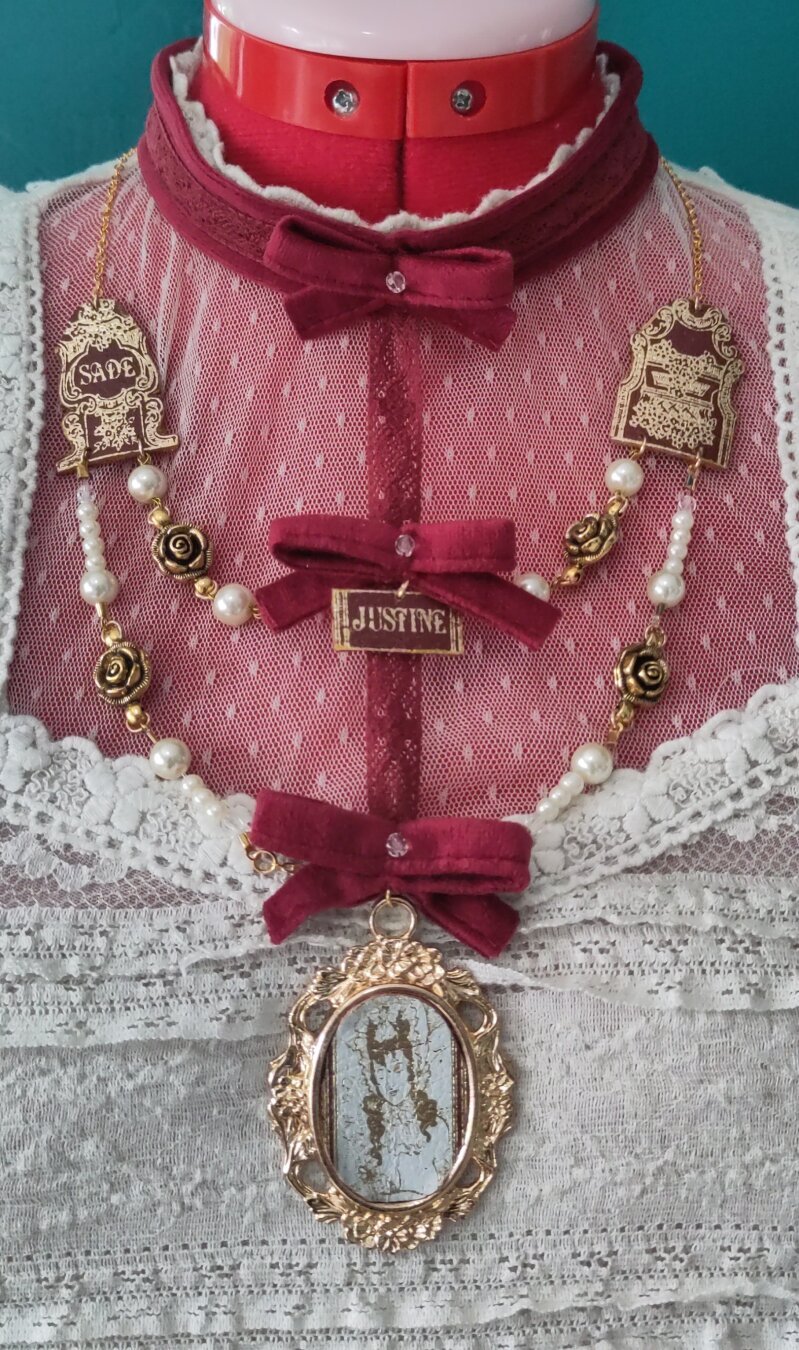 The necklace ¨Justine¨ made by MercuRea brand, consist on a red velvet chocker with a ribbon in the front, and a vertical piece of red lace, decorated with more velvet ribbons, on the side hang pieces from the spine of the book ¨Justine and the misfortunes of virtue¨ by Marquis de Sade, along with white pearls and golden rose interpieces. All of it accompanied by the golden center piece cameo featuring the artwork ¨The Billet Doux¨by Aubrey Beardsley