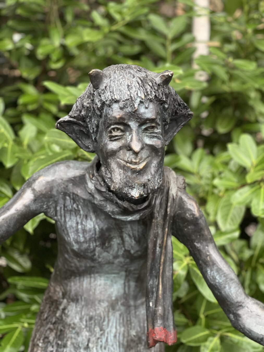 A close up photo of a statue of Mr Tumnus. He is smirking and wearing his scarf. 

It is a metal statue, with only the tip of his scarf painted with a dash of red paint 

Green foliage is the backdrop