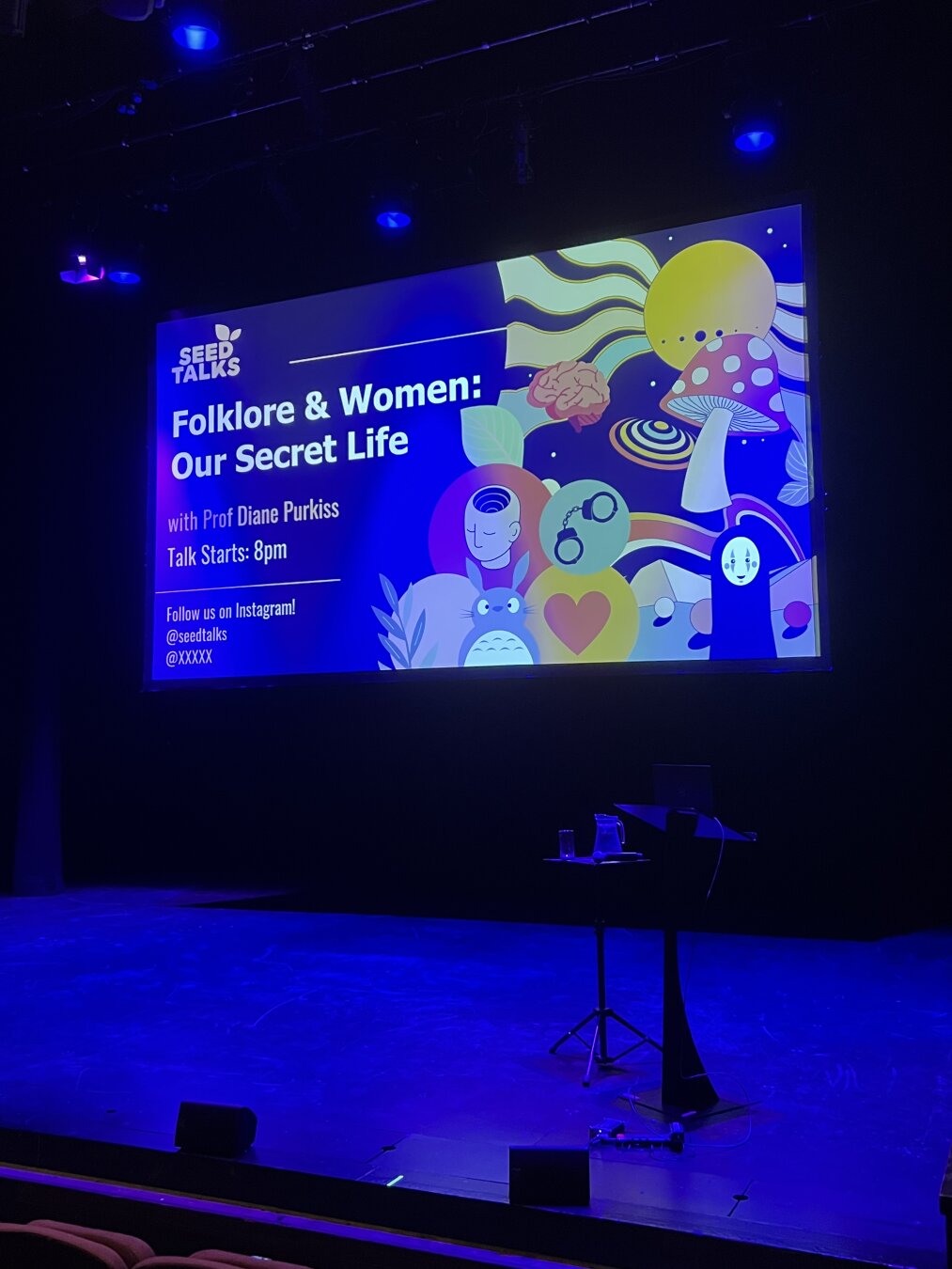 An empty stage with a screen showing the title of the talk: Folklore & Women; our secret life.