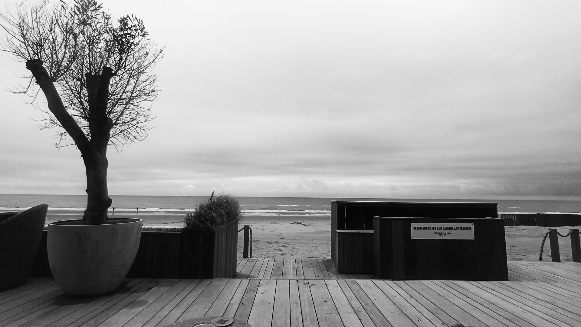 Black and white view on the North Sea between the wooden furniture on the terrace of a beach restaurant showing in the distance a couple close to the sea