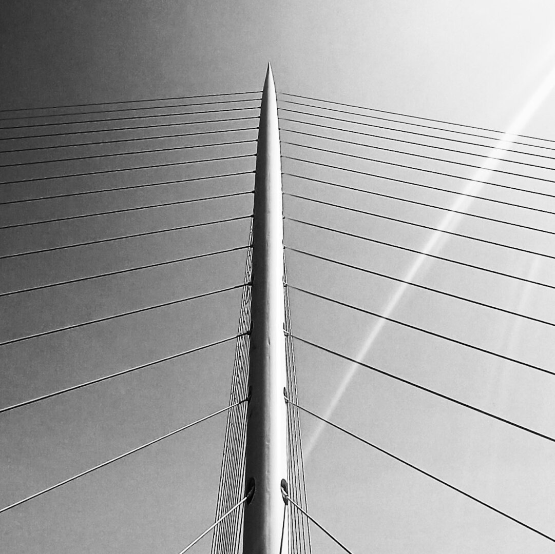 Black and white square pic showing the main pillar of a bridge with the suspension cables pointing down to the left and the right of the picture