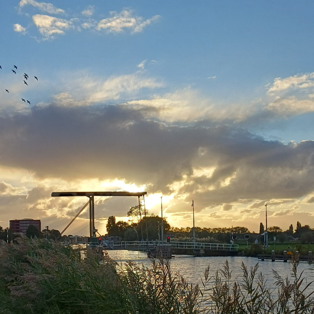 Sun setting half behind the clouds with a blue sky above and a yellow gold below, behind the cyclist's bridge across the runway near Cruquius (NL)