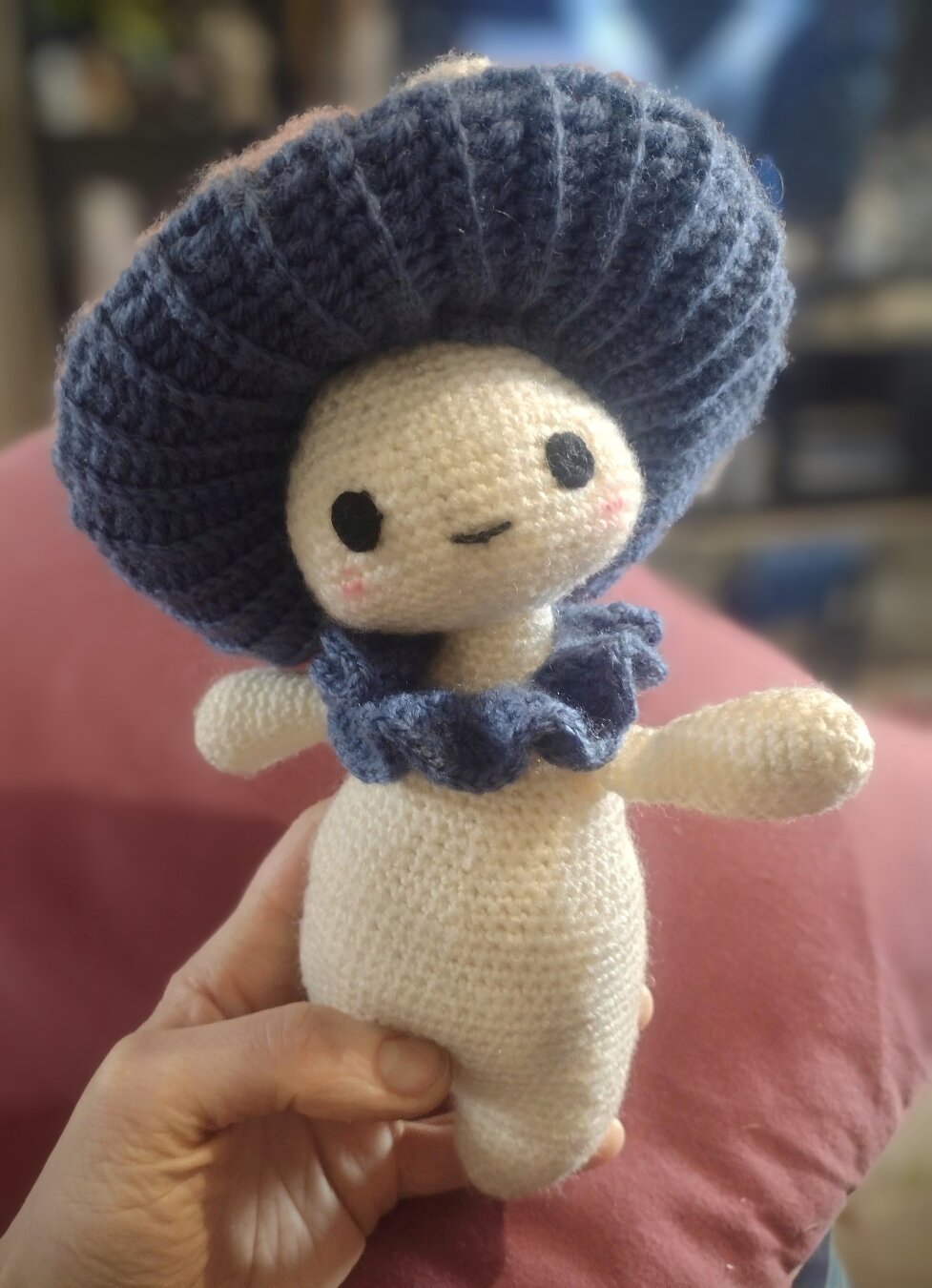 A crochet mushroom person peering up at the camera, with a slight tilt to their head.