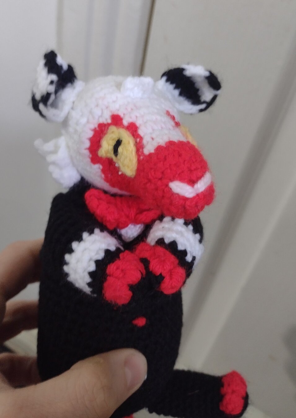 A crochet opossum in the style of Moxxie from Helluva Boss, being held upright. He has white and black striped ears, white head, and red snout. His eyes are yellow and black. His body is coloured so that it looks like he is wearing a black suit. His paws are held together in front of his body.