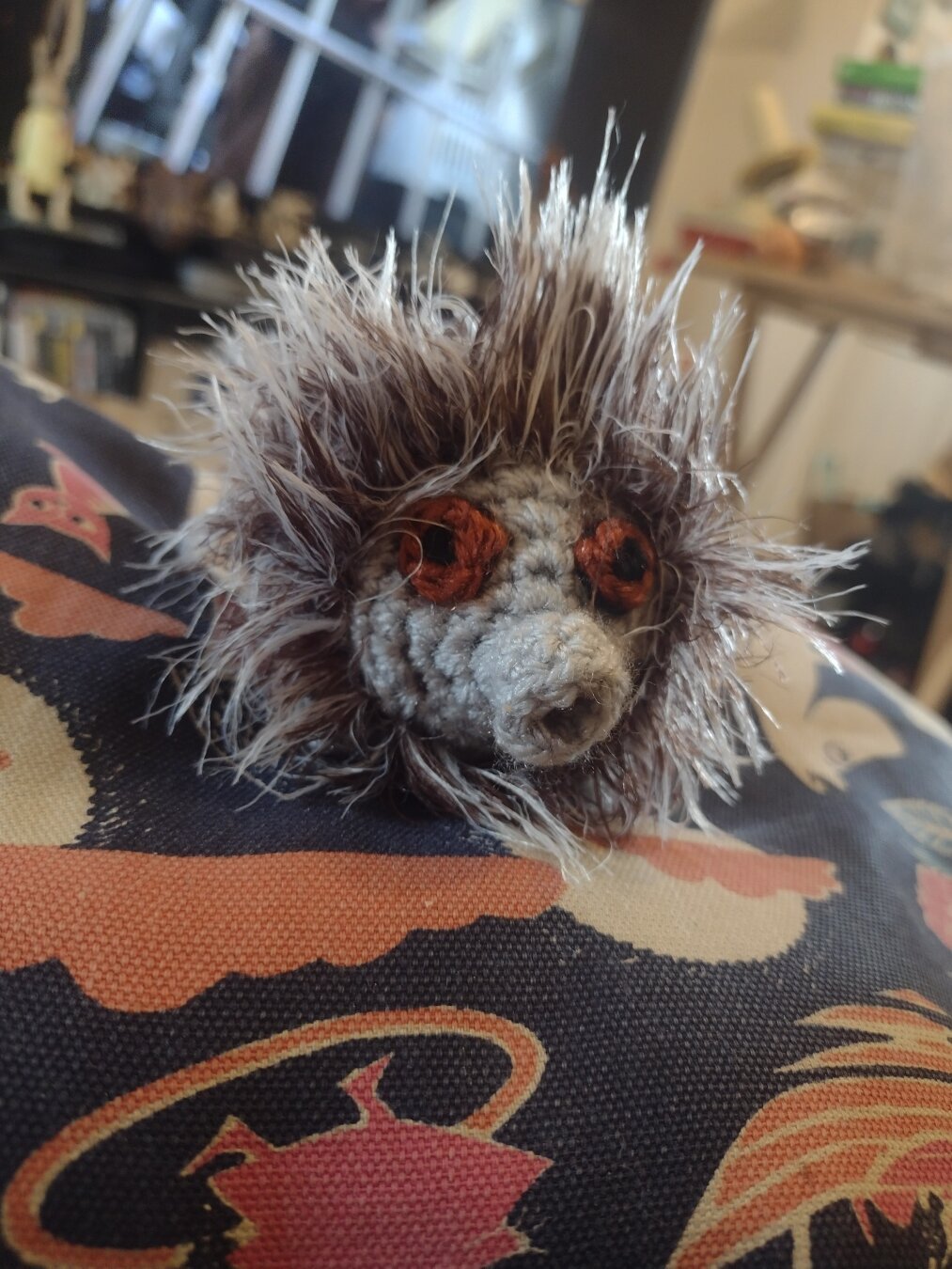 A small crochet animal. It looks somewhat similar to a hedgehog. Instead of spines it has a very fluffy grey and brown body. Its head is grey, and not fluffy. It has a very short snout that looks a bit like an elephant trunk. It has large round brown and black eyes.