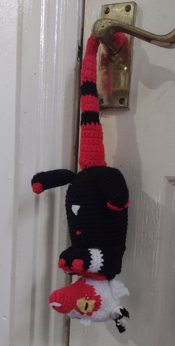 A crochet opossum in the style of Moxxie from Helluva Boss, hanging by his red and black tail on a door handle. He has white and black striped ears, white head, and red snout. His eyes are yellow and black. His body is coloured so that it looks like he is wearing a black suit. His paws are held together in front of his body.