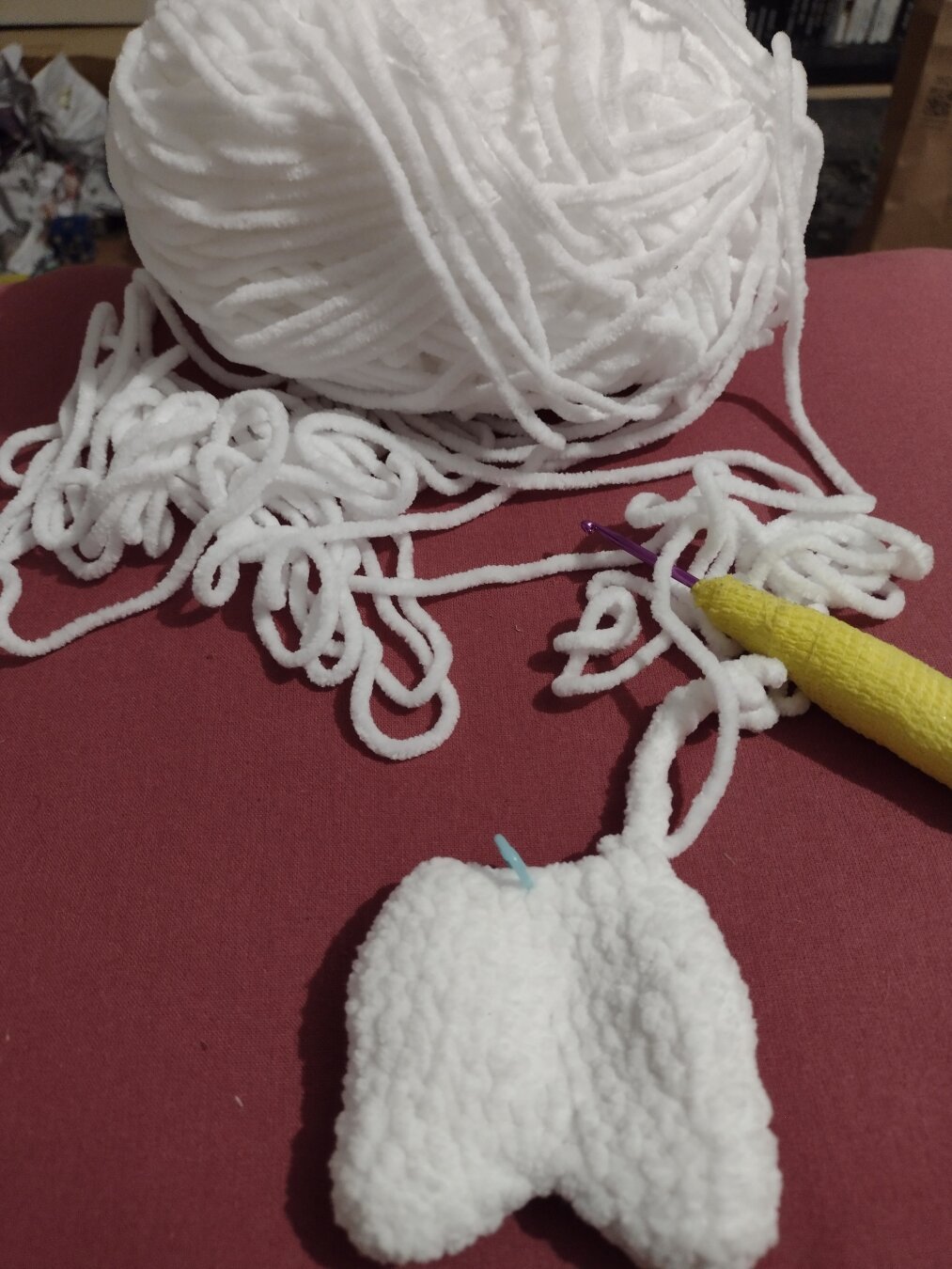 A ball of white chenille yarn, a crochet hook, and a partly made crochet amigurumi. It has two dumpy legs and half a body.