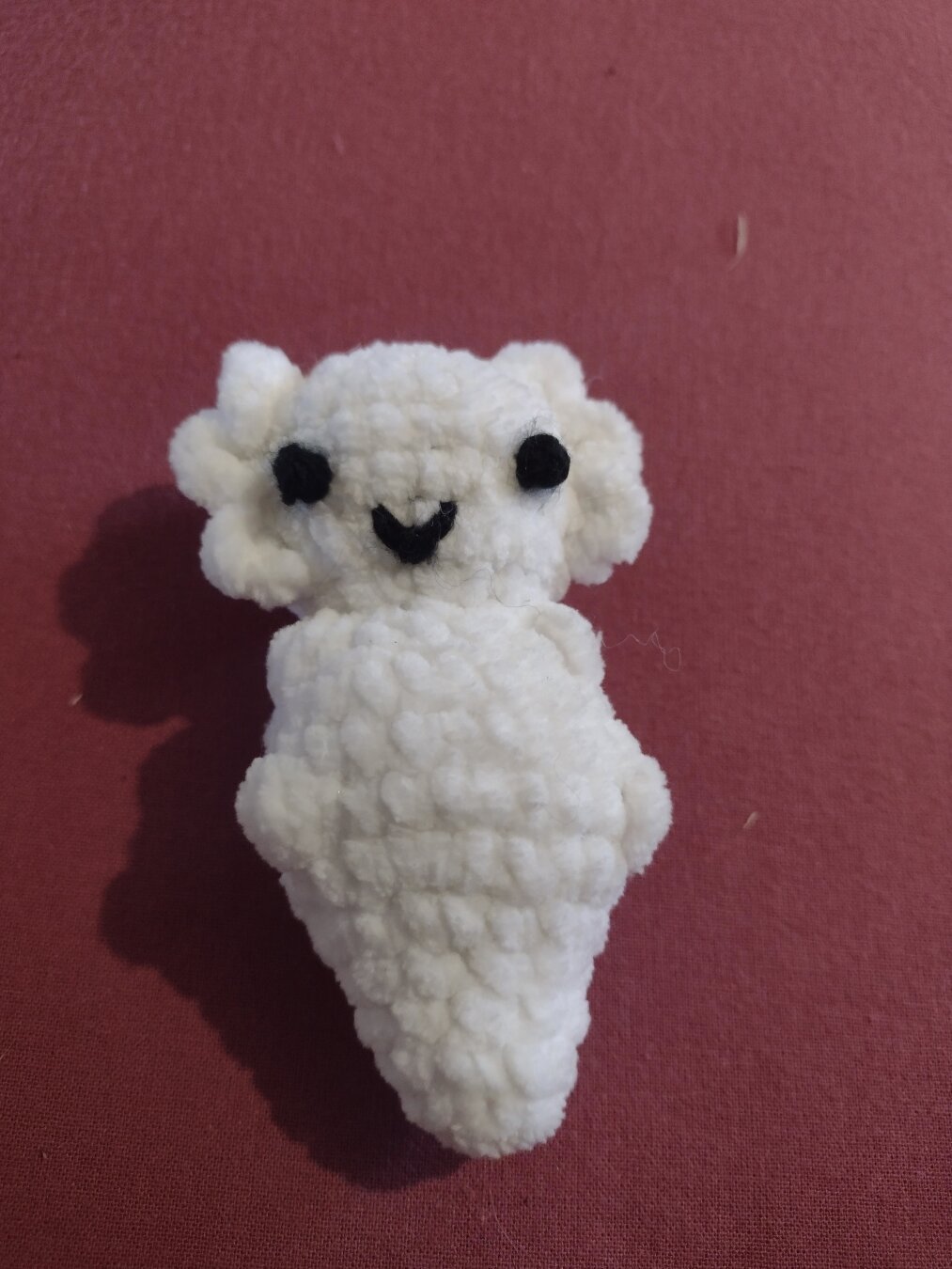 A smol white crochet axolotl. It has big black eyes and a small black mouth. It's kind of a little guy.