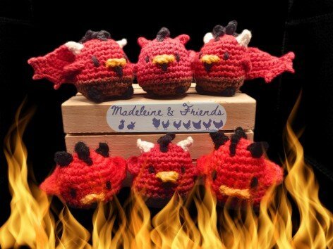 Six small crochet chickens in the style of devils. They have red upper bodies, brown lower bodies, beady black eyes, black combs and goatees under their beaks. They have horns in white, black or red, and two have extra bat wings on their back. There are flames licking the bottom of the picture. A watermark is in the centre that says "Madeleine & Friends".
