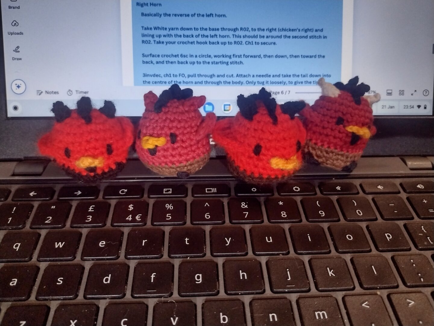 Four small, round crochet chickens in the style of little devils, sitting at various angles on a laptop keyboard. They have red upper bodies, and brown lower bodies, black combs and little black goatees under their beaks. Two have black horns, one has white horns and one has red horns.