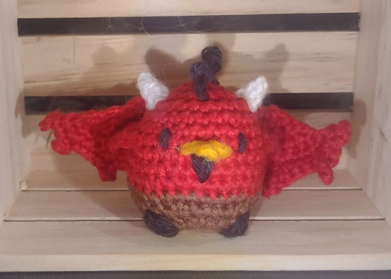 A small, round crochet chicken in the style of a devil. It has a red upper body, brown lower body, black hooves, and a black comb, and goatee under its orange beak. It has white horns. Additional to the first image, it also  has large, bat like wings on its back, as well as little dumpy chicken wings on its side.
