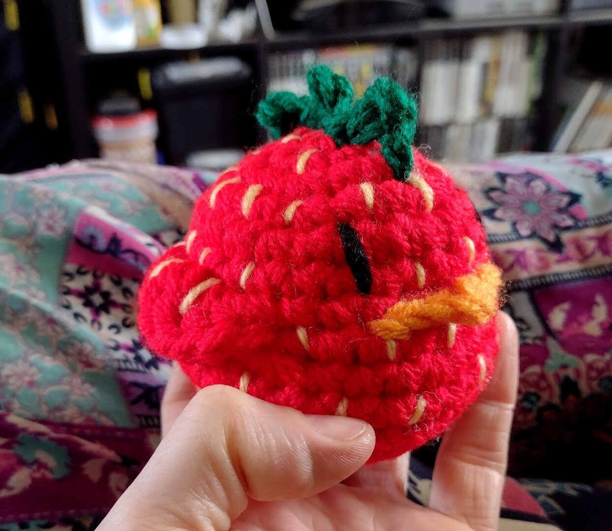 A palm sized crochet chicken. It is a little orb, with dumpy wings and no legs. It is stylised as a strawberry. It has a red body, with little yellow flecks for seeds. It's comb is green like the leaves of a strawberry plant.