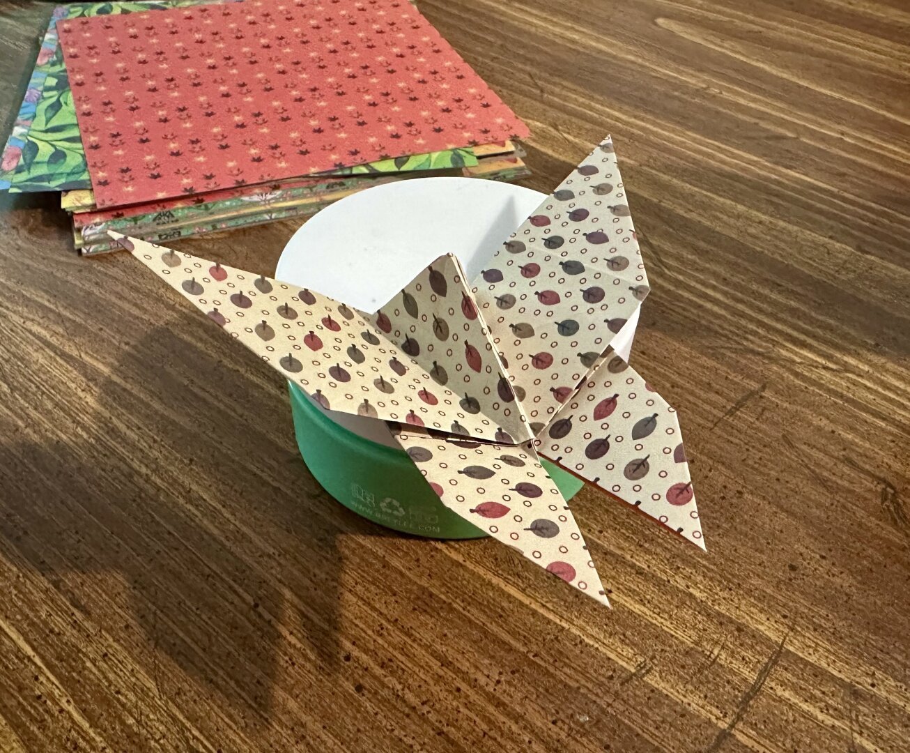 An origami butterfly made of off white colored paper with a leaf pattern. Red and green Origami paper is stacked in the background.