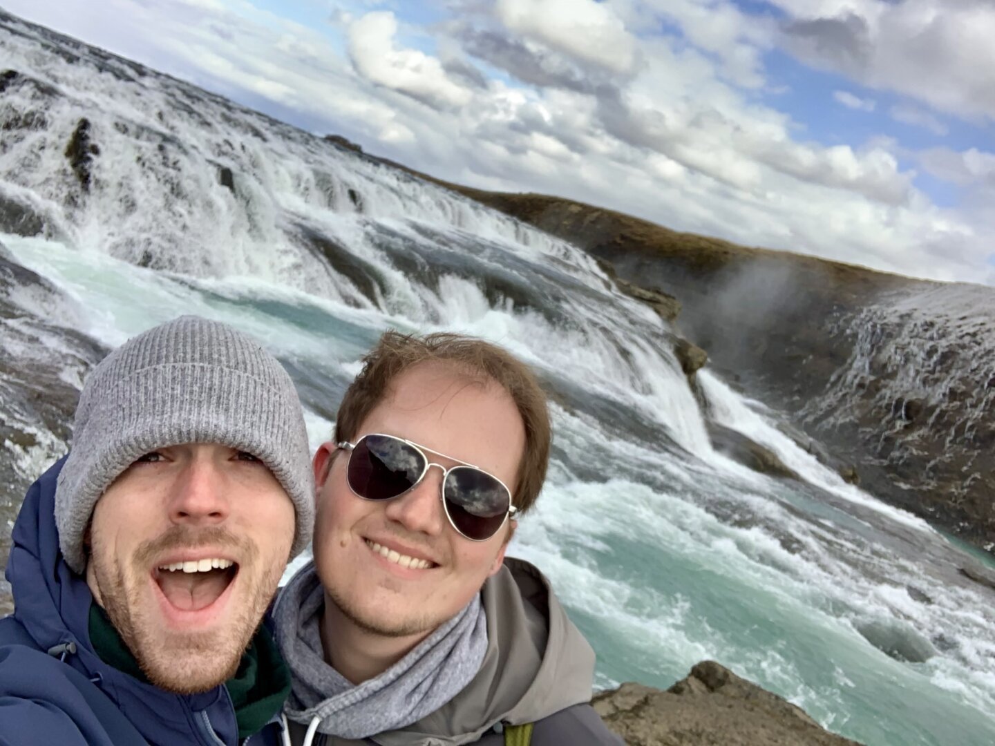 A selfie of two smiling men in front of a waterfall. The man on the left is wearing a gray beanie, the man on the right is wearing sunglasses.
