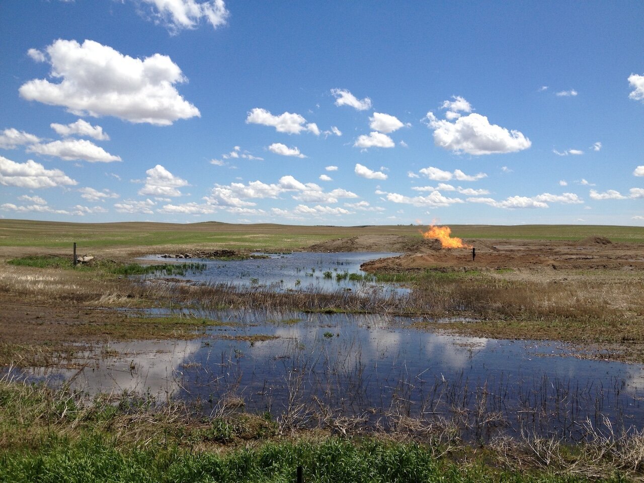 Title: Clear Skies and Fresh Water Threatened by Burning Gas

Alt Text: Photograph of a prairie landscape on a sunny day with a pond in the foreground. A few clouds drift by. A pipe is protruding from the ground, out of which is flaring burnt natural gas.

Photographer: Marshall Mayer aka @AuthenticPhoto

Date and Time: June 10, 2013 at 2:50 pm

Location: 80th Street Northwest, Wildrose, ND USA

License: CC BY-NC-ND 4.0

Caption: Driving through the American Northern Plains on a perfect late spring day, the landscape is on fire. There is so much natural gas being released in the Bakken by fracking that it can't economically be recovered! And there is no environmental protection that requires recovery. 

Hashtags: #Fracking #OilAndGas #BoomAndBust #UnnaturalDisaster #ClimateEmergency #BurnItAll #Extraction #Deregulation #Authentic #Photography

Source: https://take-note.com/note-takers/marshall--mayer/notes/clear-skies-and-clean-water-threatened-by-burning-gas/view