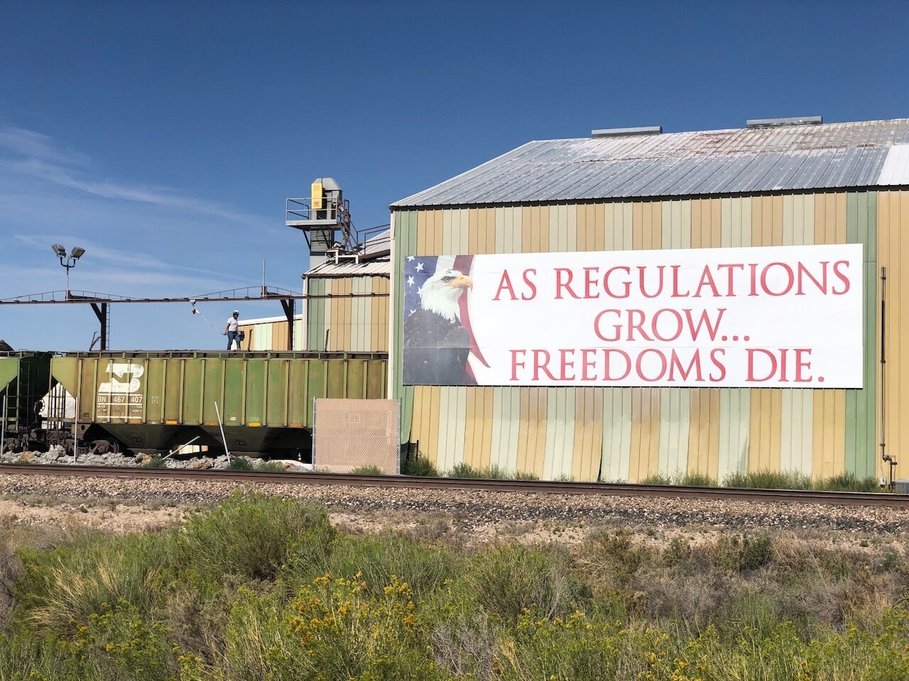 Title: Without Protections, Workers are Free to Die

Alt Text: Photograph of a worker harnessed atop a train car being loaded. The train is emerging from a warehouse, upon which is a huge sign, 