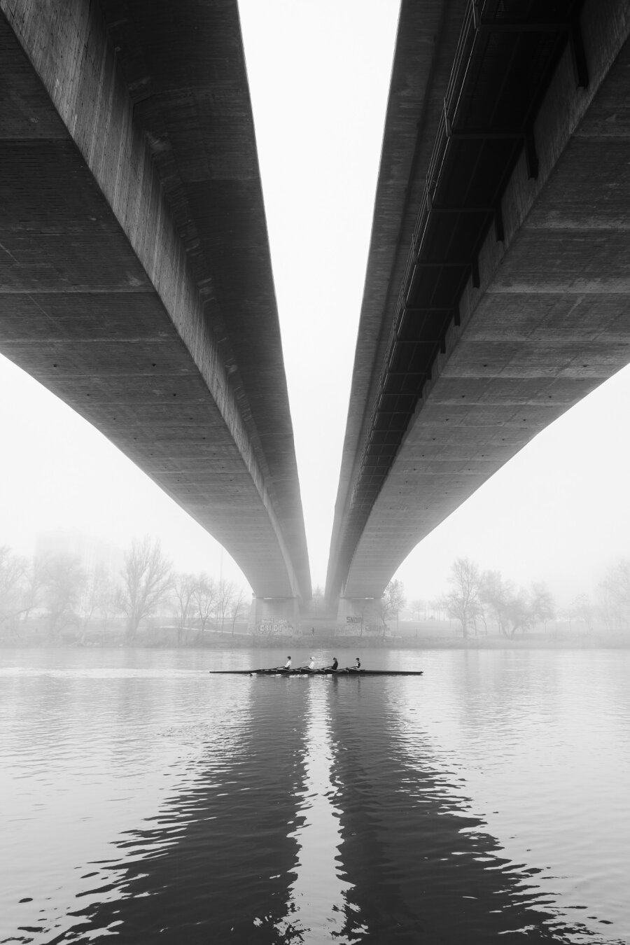 A black-and-white photograph captures a symmetrical view of two massive concrete bridges converging in the distance, creating a striking V-shape. Beneath them, a rowing team glides across the calm river, their boat and reflections forming a horizontal contrast to the towering vertical structures. The water’s surface is gently rippled, mirroring the bridges and the rowers. A layer of mist softens the background, where bare trees and faint outlines of buildings are visible along the riverbank. The composition balances the rigid geometry of the man-made structures with the fluidity of water and the human presence, evoking a sense of scale, symmetry, and quiet movement.