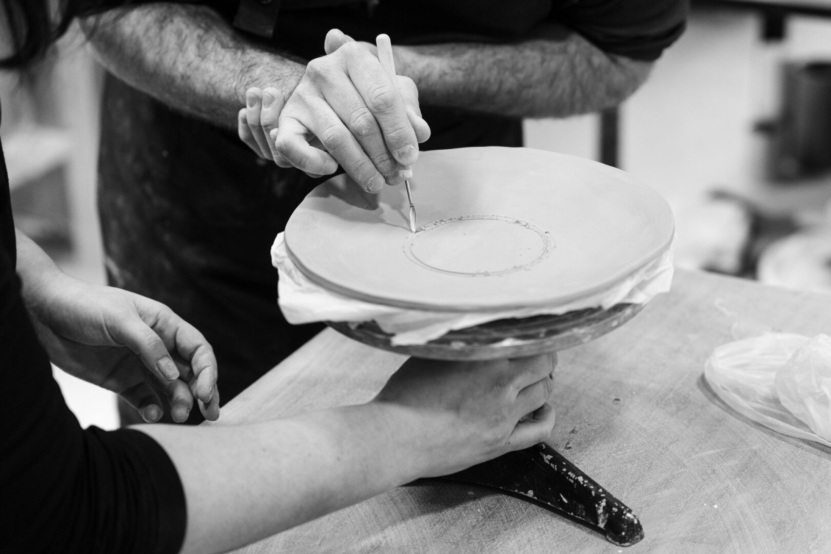 A black and white photograph captures a close-up of two pairs of hands working together on a ceramic piece. The hands belong to an instructor and a student, engaged in a pottery lesson. The instructor's hands gently guide the student's as they carve a circular design onto the smooth clay surface of a plate, which rests on a rotating stand. The student's left hand steadies the stand while their right hand follows the mentor's lead. The texture of the clay, the fine details of the carving tool, and the contrast between light and shadow emphasize the craftsmanship and learning process. The background, slightly blurred, suggests a workshop setting with tools and materials scattered around.