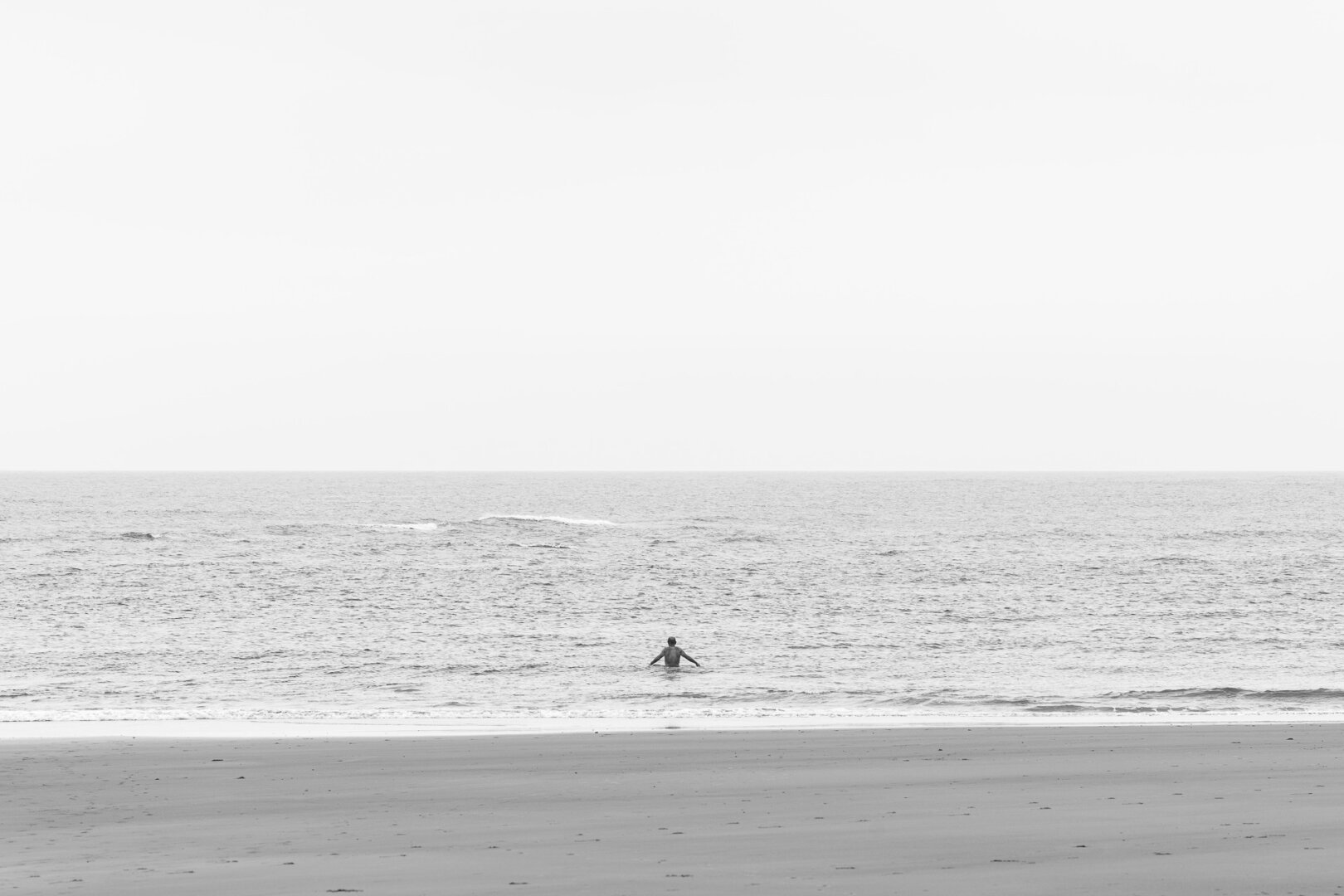 A black and white photograph of a solitary person standing in the ocean, facing the horizon. The composition is minimalistic, with the vast sky occupying most of the frame, blending seamlessly with the calm sea. The figure, positioned near the center, appears small against the expanse of water and sky, evoking a sense of solitude and introspection. Gentle waves ripple across the ocean's surface, contrasting with the smooth, empty shoreline in the foreground. The high-key exposure enhances the ethereal and serene atmosphere, emphasizing the contrast between the dark water and the luminous sky.