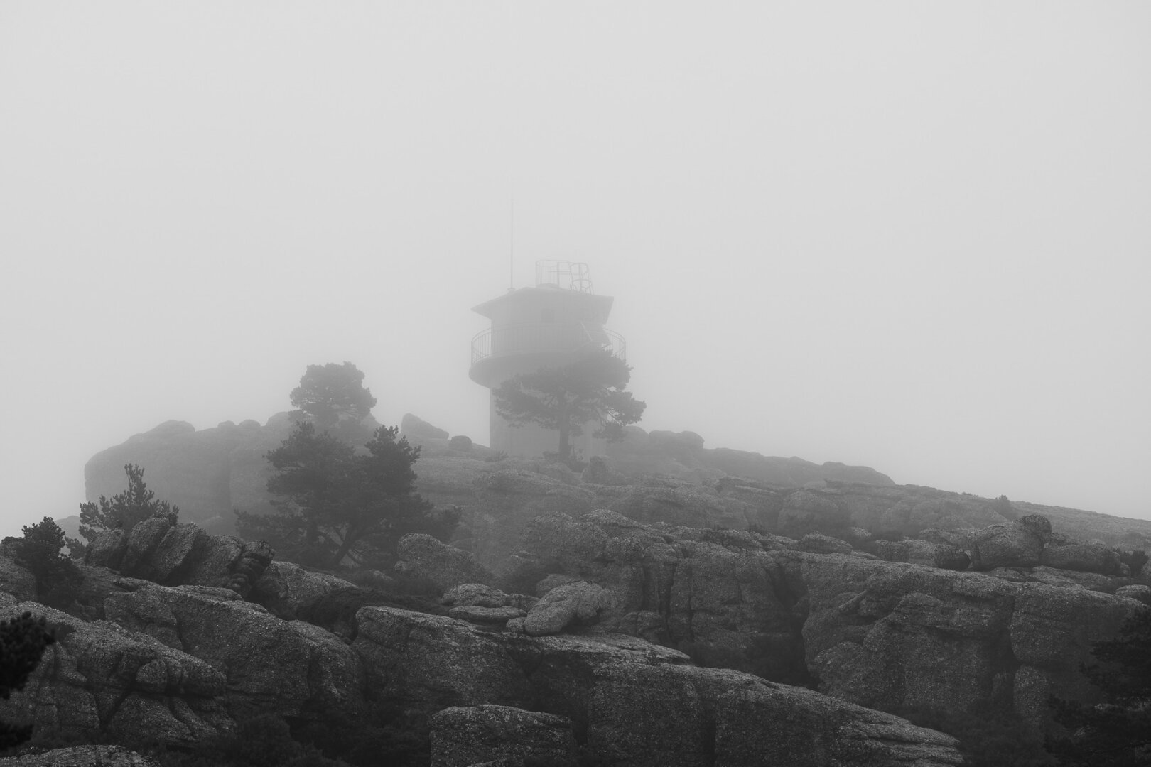 A black-and-white photograph of a solitary watchtower partially obscured by dense fog, standing atop rugged, rocky terrain. The scene is enveloped in mist, creating a mysterious and haunting atmosphere. Dark pine trees emerge through the fog, adding to the eerie, otherworldly feel. The smooth, rounded boulders and jagged rocks contrast with the soft, diffused light, emphasizing the bleakness and isolation of the landscape. The fog blurs the line between the land and sky, giving the impression of a timeless, suspended moment.