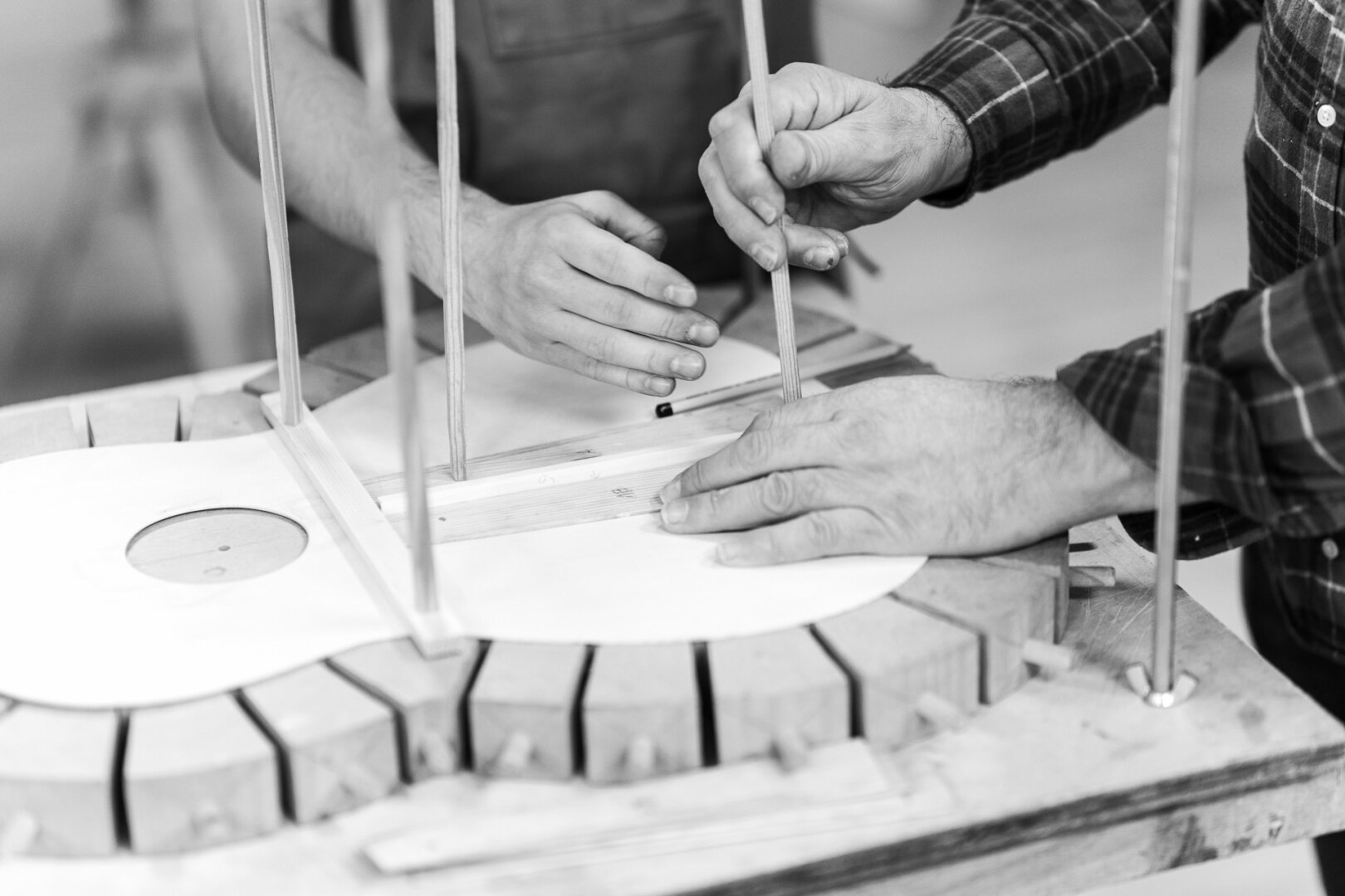 A black and white photograph captures the hands of two artisans working together on the construction of a guitar. The image focuses on their precise movements as they carefully position wooden braces on the instrument’s top, which is secured by a traditional clamping system. The contrast between the hands—one appearing more experienced and the other younger—suggests a moment of mentorship and craftsmanship. The surrounding tools and structured workspace hint at the meticulous process involved in luthiery, the art of guitar making.