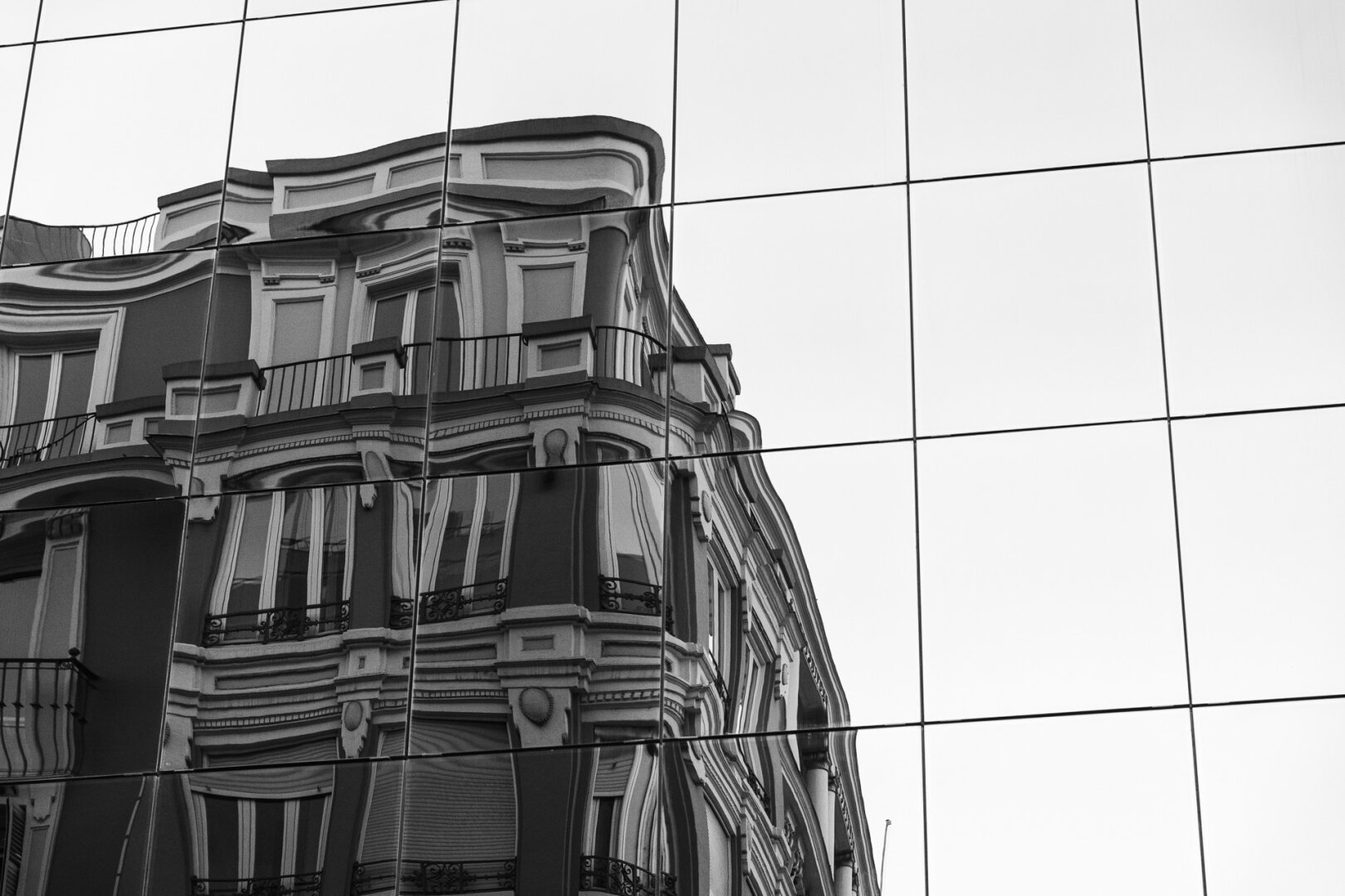 A black and white photograph captures the distorted reflection of a historic building on the glass facade of a modern structure. The reflection is fragmented by the grid of windowpanes, creating a surreal, wavy effect that bends the building’s ornate balconies, windows, and decorative elements. The juxtaposition of old and new architecture adds a sense of contrast and abstraction, emphasizing the interplay between past and present in the urban landscape.